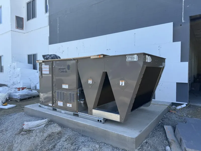 A large grey commercial HVAC unit sits on a concrete pad at a construction site