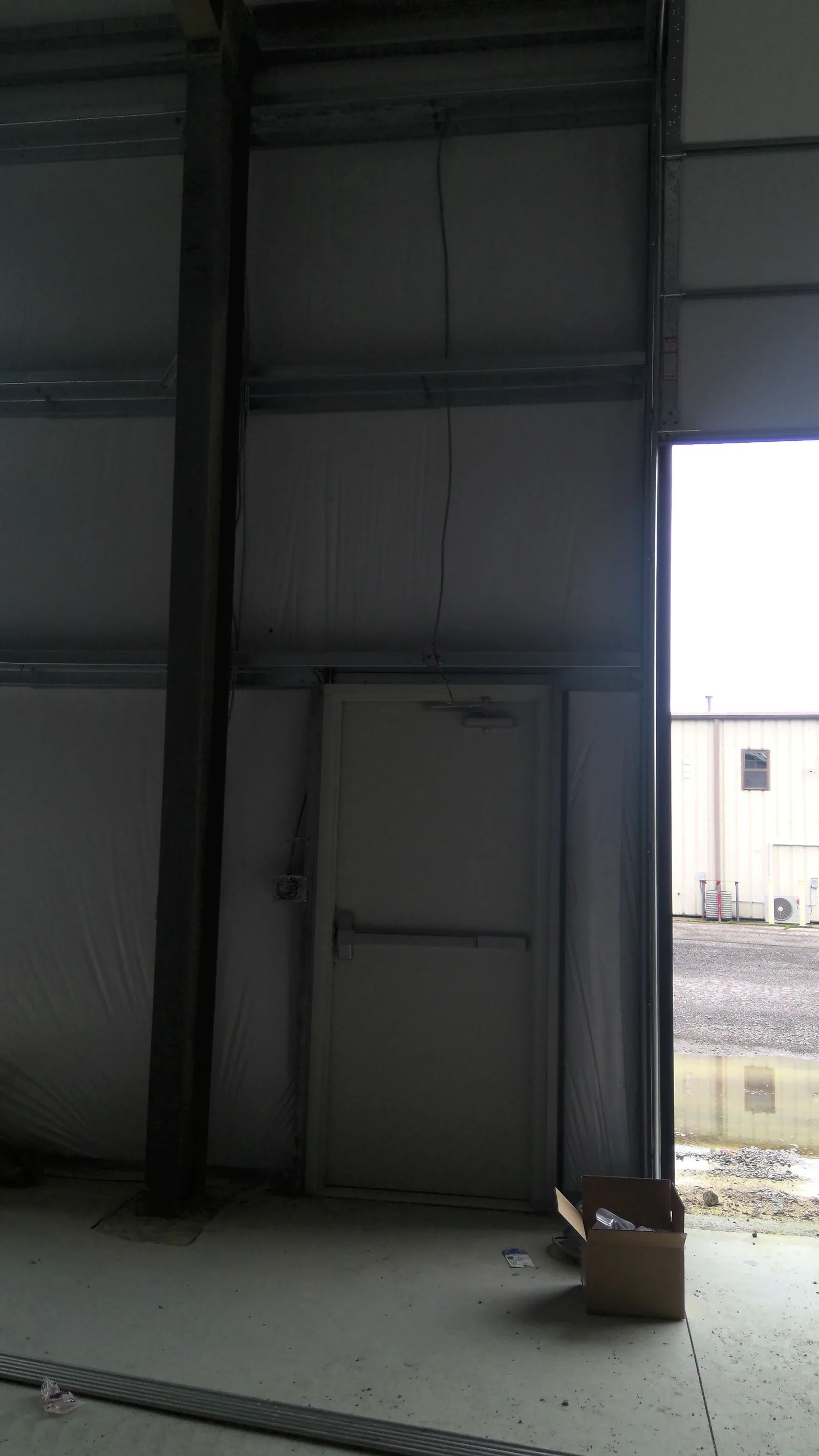 An interior warehouse wall showing a metal exit door and wiring next to an open bay