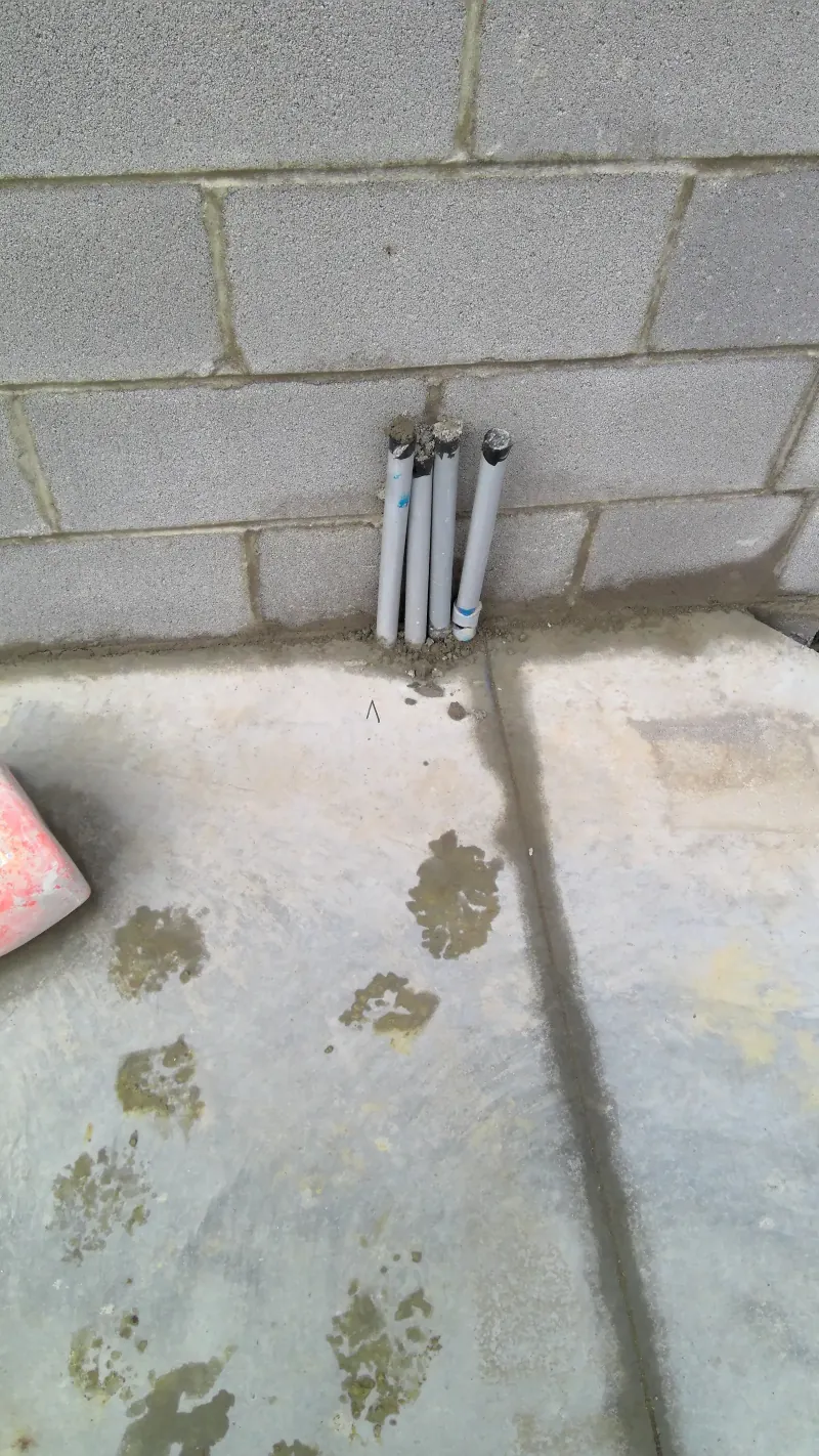 Four grey electrical conduits and pipes come through a concrete floor next to a block wall